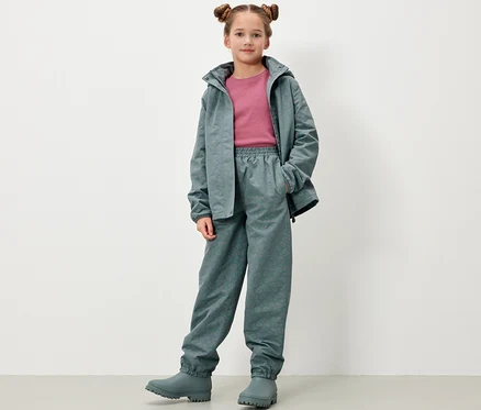 Mädchen posiert in Kinder-Regenhose mit reflektierenden Elementen, Jacke und Gummistiefeln in Graugrün.