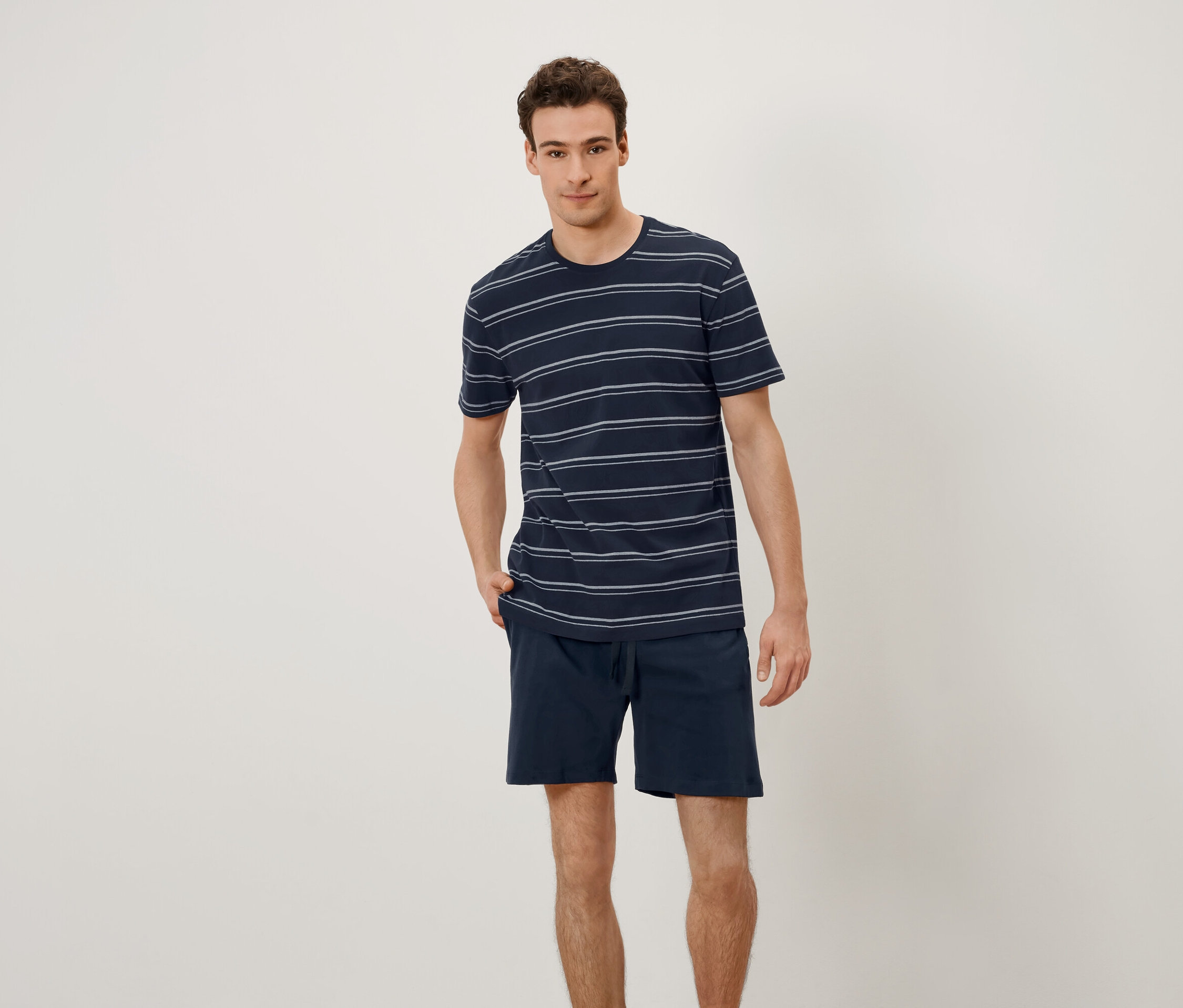 Ein Mann steht und hat seine Hände in den Hosentaschen. Er trägt ein T-Shirt mit horizontalen Streifen und Shorts.