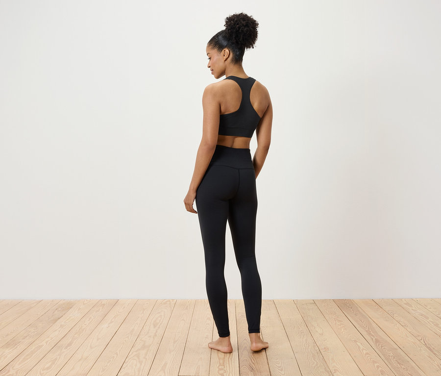 Frau in schwarzem Sport-BH und Leggings steht auf Holzboden, mit dem Rücken zur Kamera.