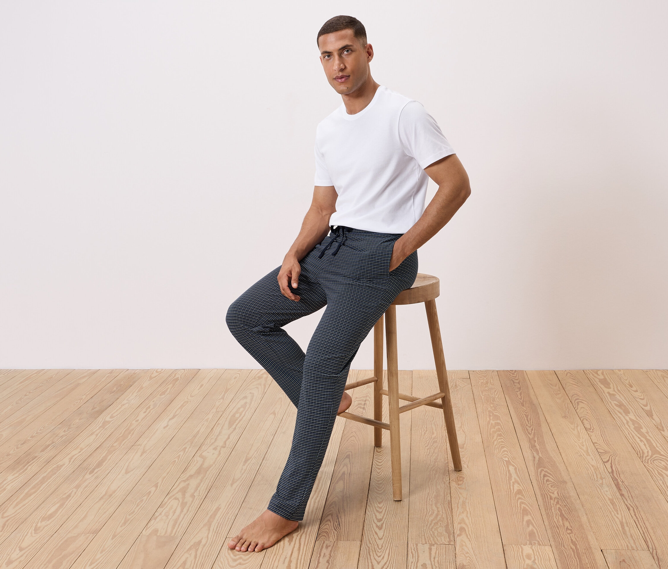 Mann sitzt auf einem Hocker, trägt ein weißes T-Shirt und gemusterte Hosen.