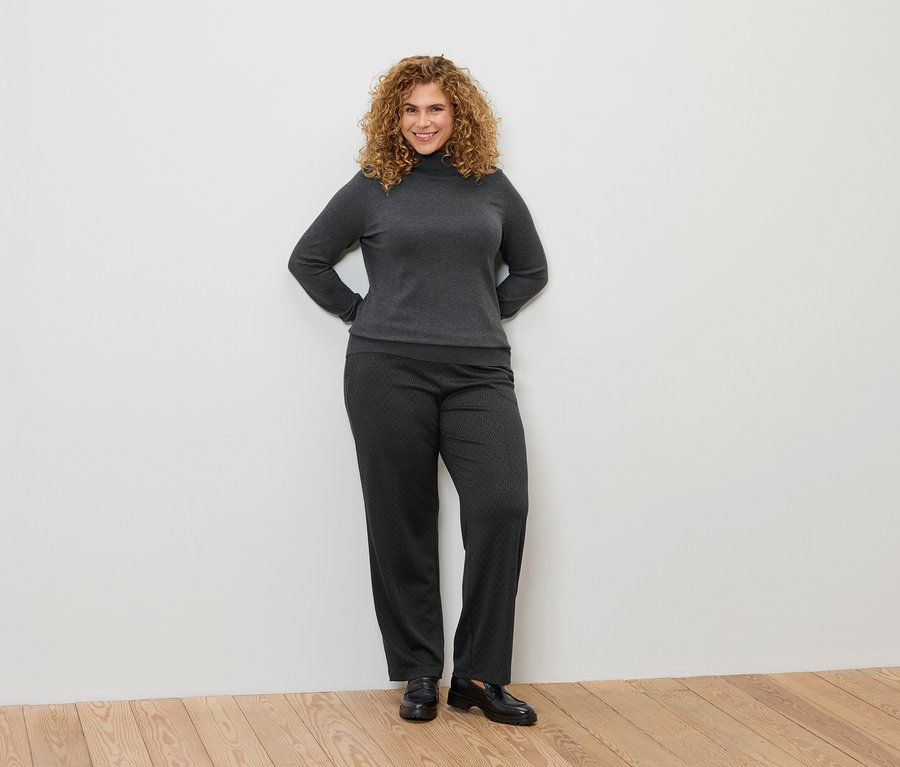 Frau mit lockigem Haar posiert in einem grauen Rollkragenpullover und gemusterten Hosen.