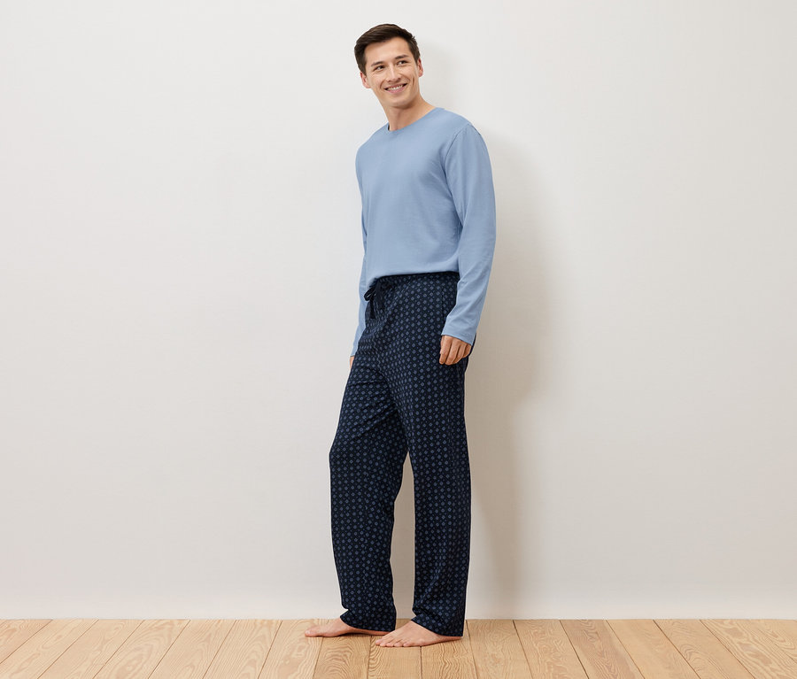 Ein Mann steht barfuß vor einer weißen Wand und trägt einen hellblauen Pullover und eine dunkelblaue Pyjamahose mit Punktmuster.