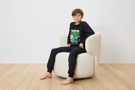 Ein Junge sitzt barfuß in einem cremefarbenen Sessel und trägt einen schwarzen Minecraft-Pyjama.