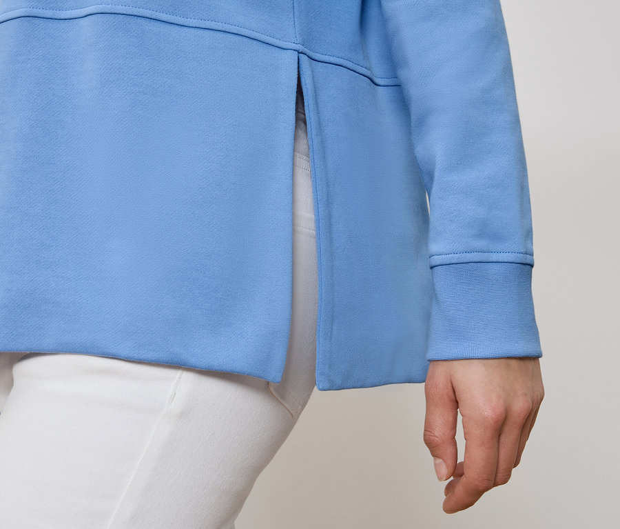 Person trägt hellblaues Sweatshirt mit Seitenschlitz und weiße Hose.