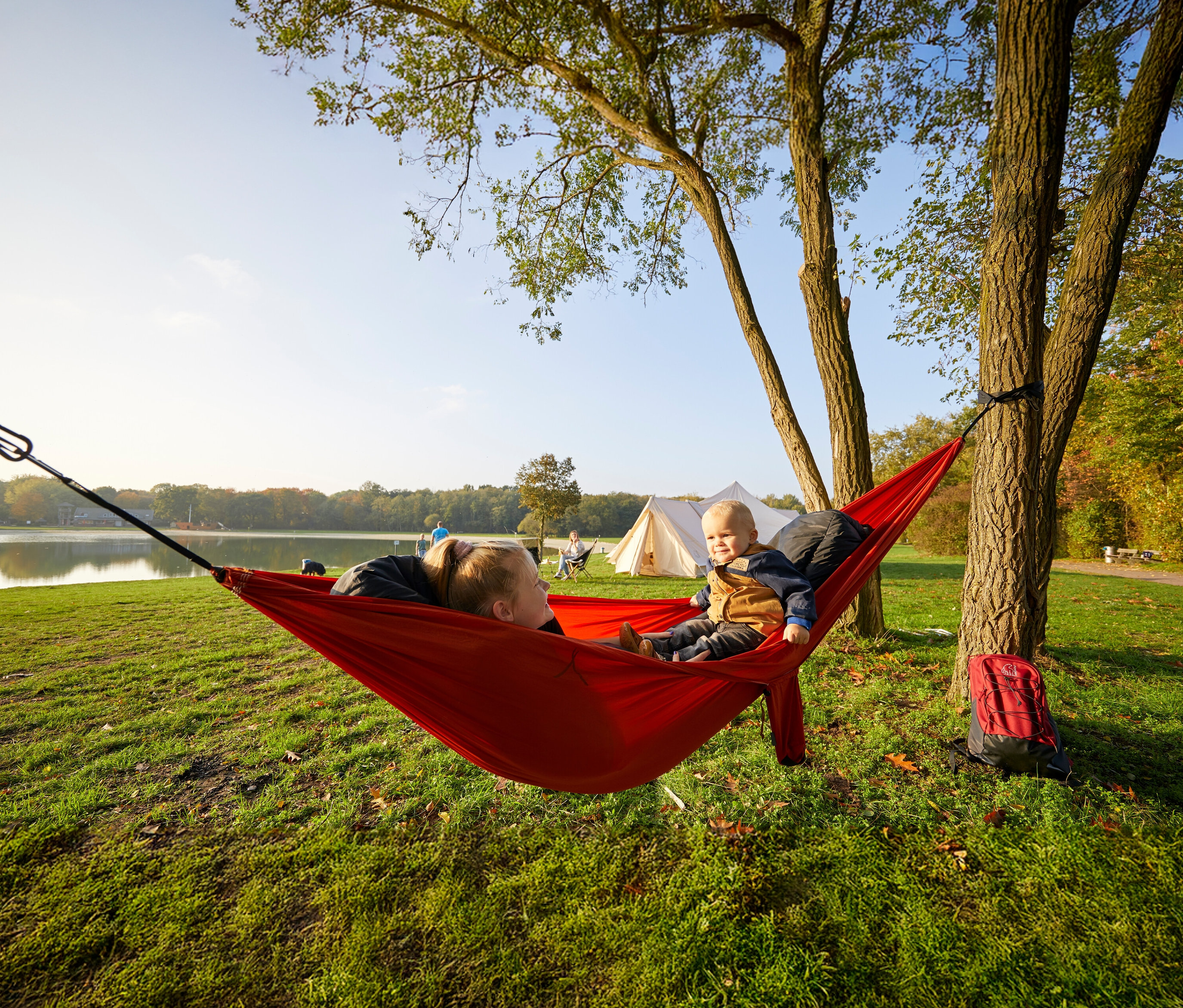 Eine Frau und ein Kleinkind liegen in einer roten Grand Canyon Doppel-Hängematte »Bass Hammock«, die zwischen zwei Bäumen aufgespannt ist.