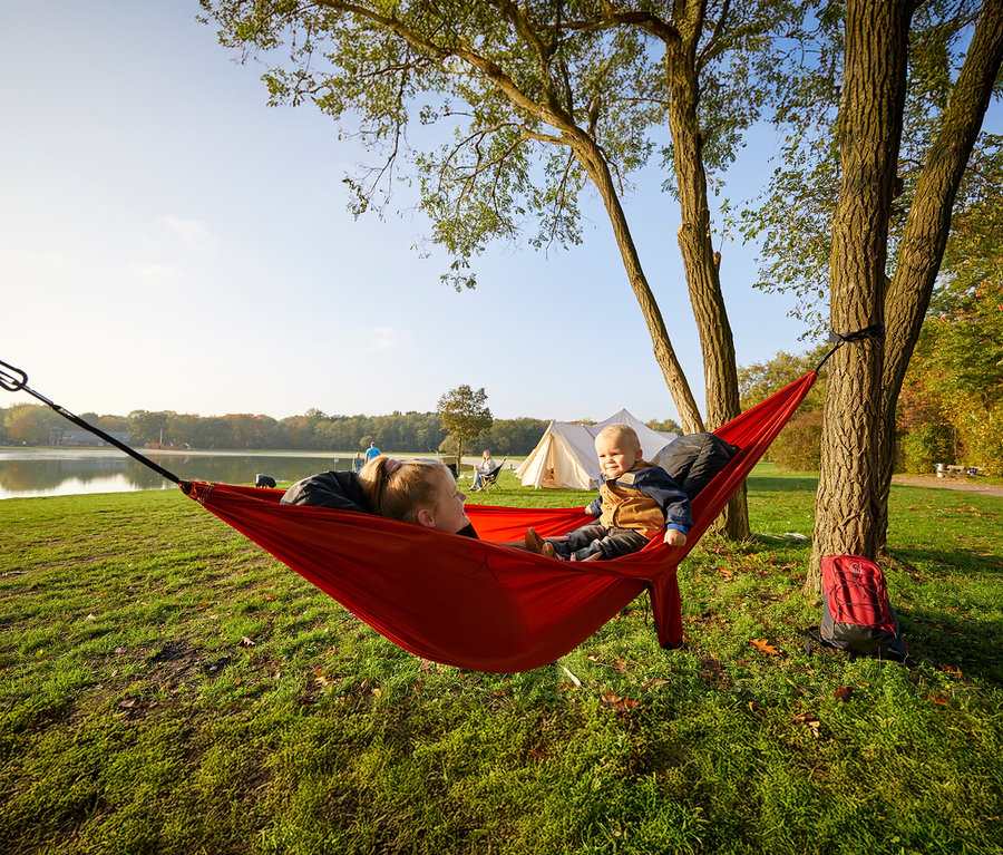 Eine Frau und ein Kleinkind liegen in einer roten Grand Canyon Doppel-Hängematte »Bass Hammock«, die zwischen zwei Bäumen aufgespannt ist.