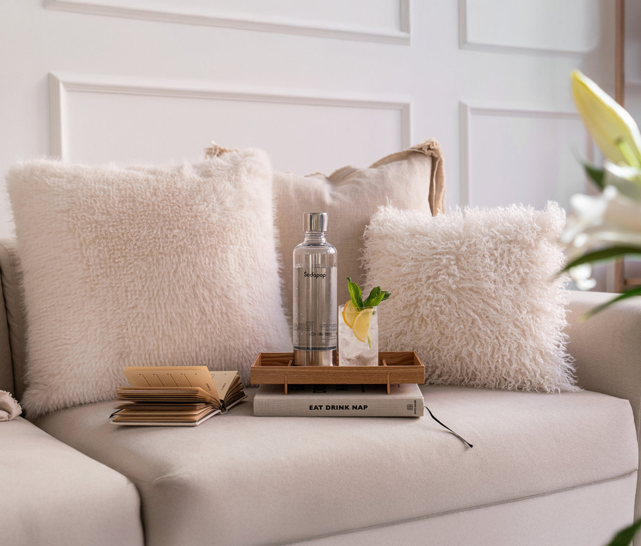 Auf einem beigen Sofa stehen zwischen Kissen ein Sodapop Wassersprudler Cooper Pro in Weiß, inkl. 5 Flaschen, ein Tablett mit einem Getränk und ein Buch.