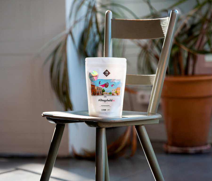 Eine Packung Alltagsheld Kaffee sitzt auf einem grünen Stuhl vor einer Pflanze.