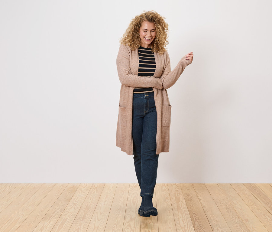 Frau mit lockigem Haar trägt einen langen, hellbraunen Cardigan über einem schwarz-weiß gestreiften Oberteil und dunkle Jeans.