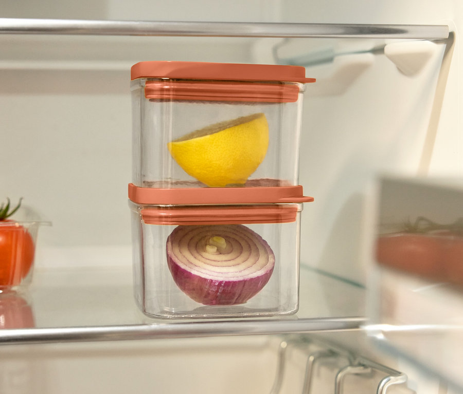 Im Kühlschrank stehen zwei durchsichtige Behälter mit orangefarbenen Deckeln. In einem Behälter befindet sich eine halbe Zitrone, im anderen eine halbe rote Zwiebel. Links davon eine Schale mit Tomaten.