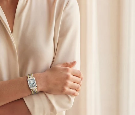 Frau mit verschränkten Armen trägt eine silberne und goldene rechteckige Armbanduhr vor einem beigen Vorhang.