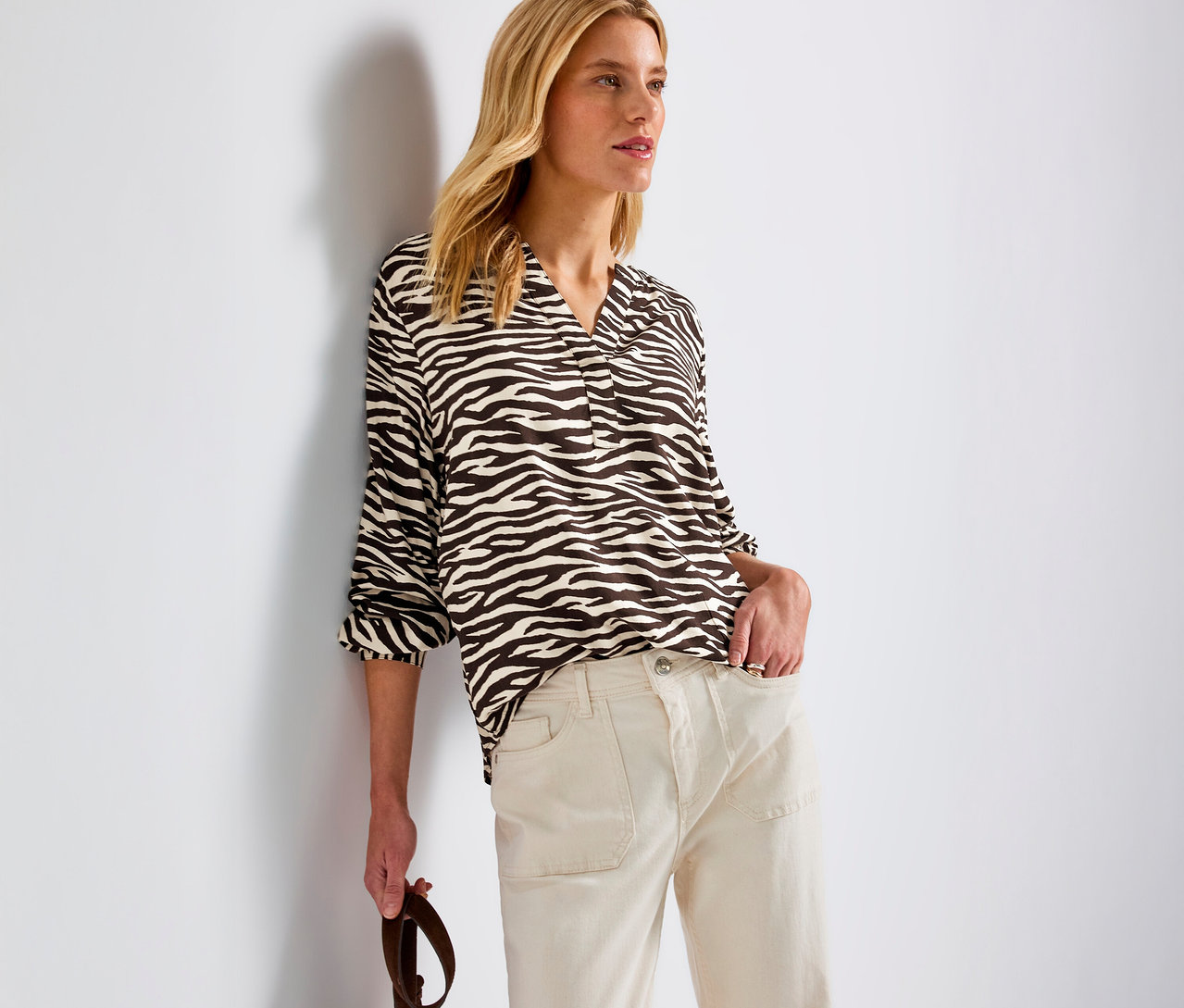 Eine blonde Frau lehnt an einer Wand und trägt eine Street One Bluse mit Zebramuster und helle Hosen.