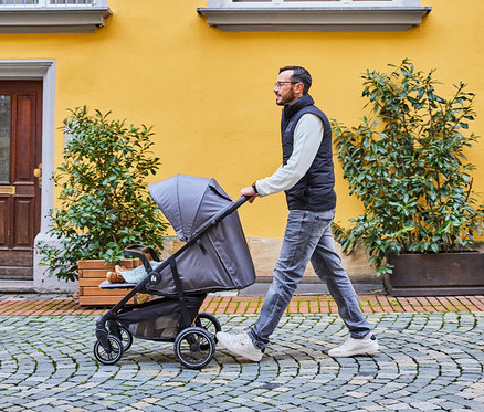 Mann schiebt einen grauen Kinderwagen auf einem gepflasterten Weg vor einem gelben Gebäude.