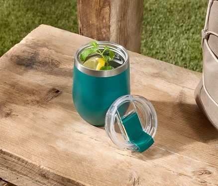 Ein grüner, isolierter Becher mit einem Getränk mit Zitrone und Minze steht mit seinem Deckel auf einem Holztisch neben einer beigen Tasche.