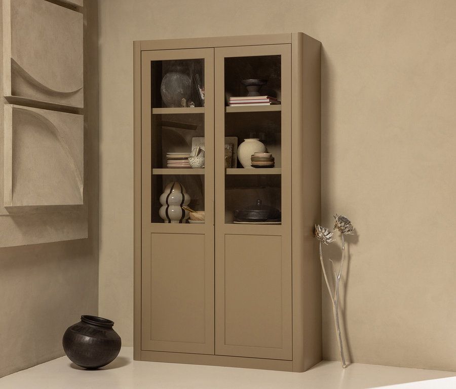 Beiger Schrank mit Glasfronten, gefüllt mit Vasen und Geschirr, neben einer dunklen, bauchigen Vase und Trockenblumen.