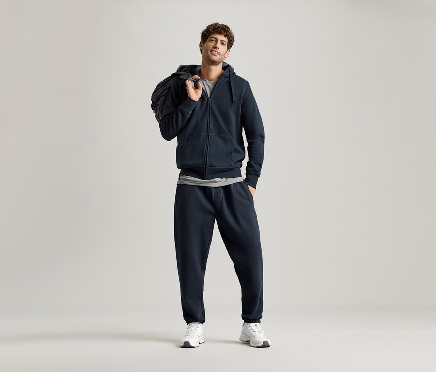 Mann in marineblauer Kapuzenjacke und marineblauer Jogginghose, mit einer Tasche über der Schulter.