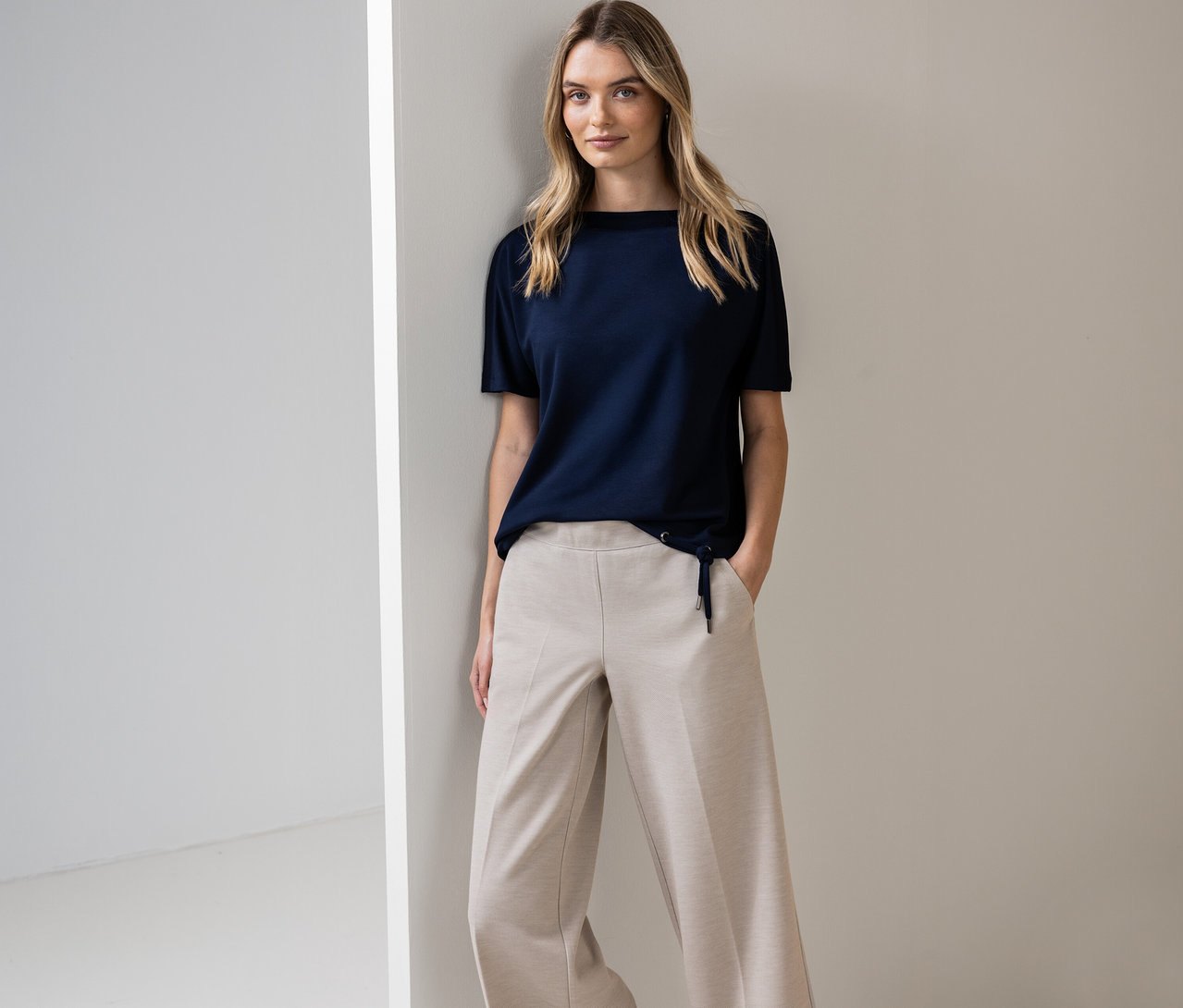 Frau posiert in marineblauem Street One Kurzarmshirt und beigen Hosen an einer Wand.