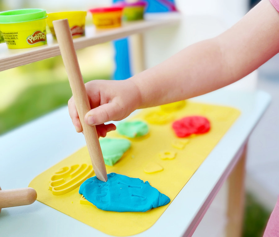 Ein Kind formt blaue Play-Doh-Knete mit einem Holzwerkzeug auf einer gelben Matte. Mehrfarbige Knetbecher stehen im Hintergrund.