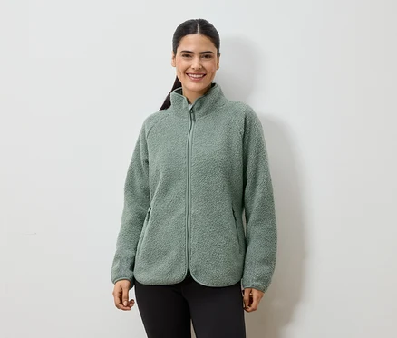 Eine lächelnde Frau mit Pferdeschwanz trägt eine grüne Fleecejacke mit Reißverschluss und schwarze Leggings vor einem weißen Hintergrund.