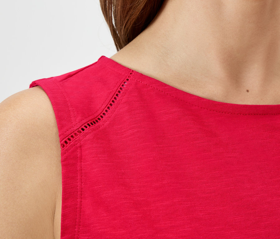 Nahaufnahme einer Frau, die ein rotes, ärmelloses Oberteil mit Lochmuster an den Schultern trägt.