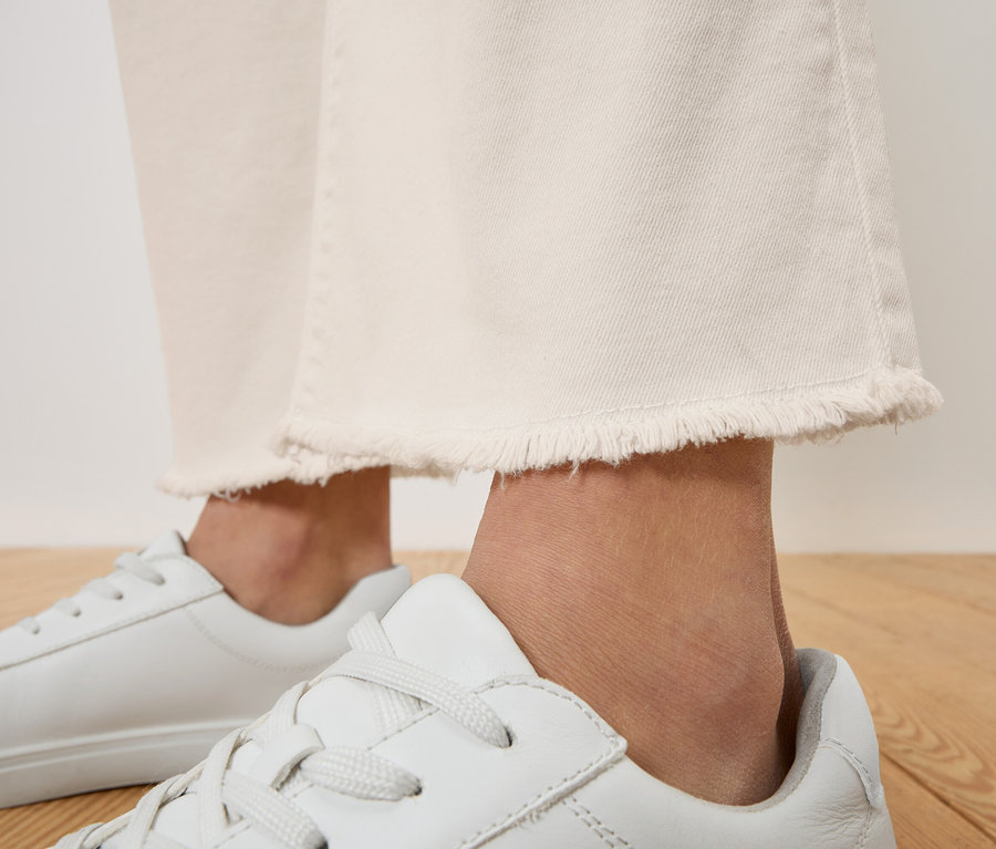 Nahaufnahme von einer Person, die weiße Jeans mit Fransen und weiße Turnschuhe trägt.