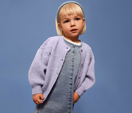 Mädchen mit blonden Haaren trägt eine helllila NAME IT Kinder-Strickjacke über einem Jeanskleid vor blauem Hintergrund.