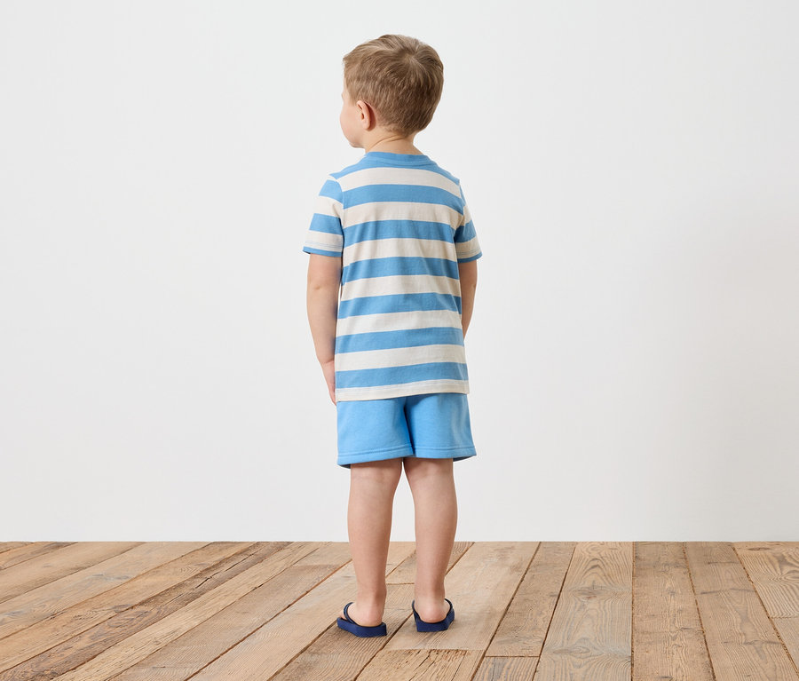 Ein Junge steht mit dem Rücken zur Kamera, er trägt ein gestreiftes T-Shirt und blaue Shorts.