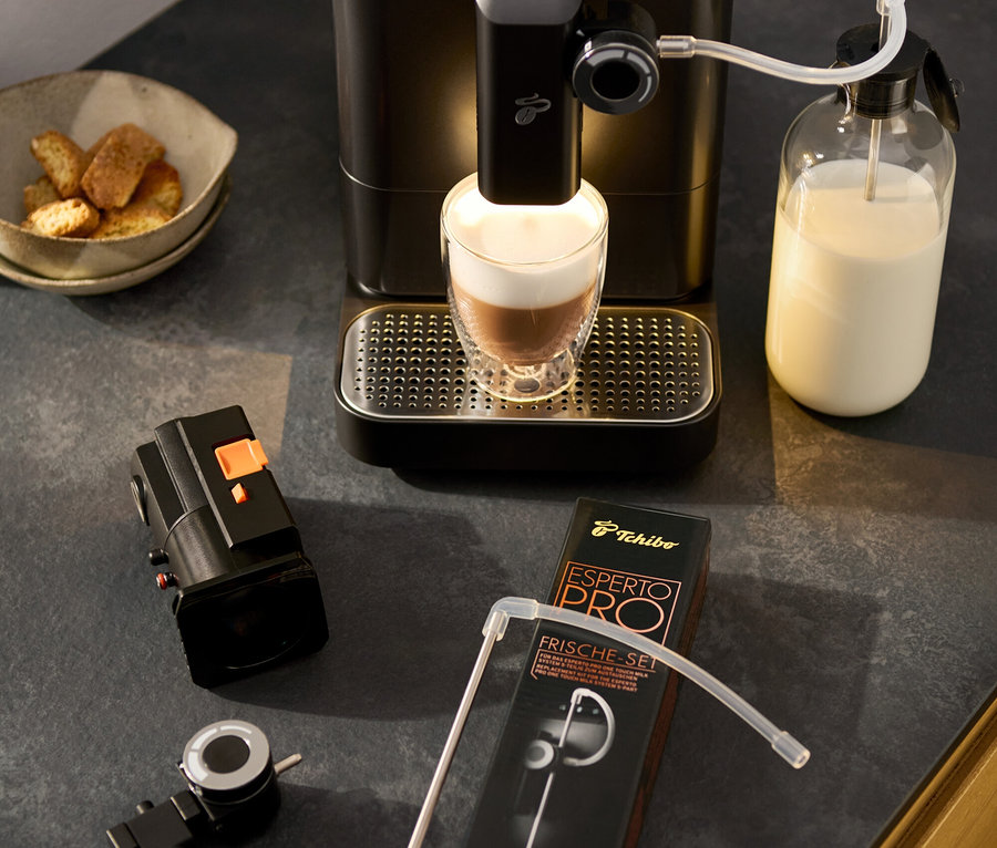 Der »Esperto 2 Milk« Kaffeevollautomat, Granite Black bereitet einen Latte Macchiato zu. Daneben stehen ein Milchschaumdrehregler, eine Schale mit Gebäck und eine Packung eines Zubehörsets.