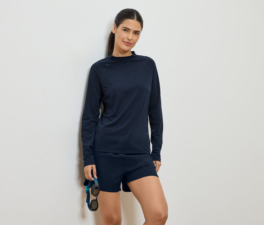Frau in marineblauem, langärmligem Oberteil und Shorts, lehnt an einer Wand und hält eine Schwimmbrille.