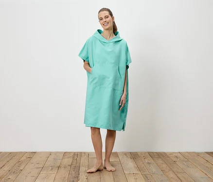 Eine Frau steht barfuß auf einem Holzboden und trägt einen türkisfarbenen Badeponcho mit Kapuze und Tasche.