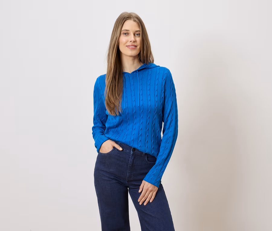 Frau mit langen Haaren posiert in blauer Strickjacke mit Kapuze und dunkler Jeans vor hellem Hintergrund.