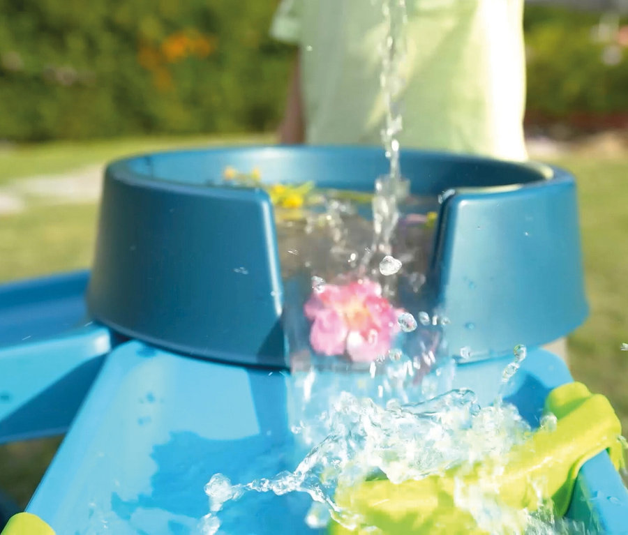 Wasser fließt in die blaue AquaPlay »Mudway STREAM« Matschbahn, in der gelbe und pinke Blumen schwimmen.