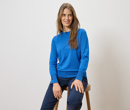 Frau mit langen Haaren sitzt auf einem Hocker und trägt einen blauen Pullover und eine blaue Hose.