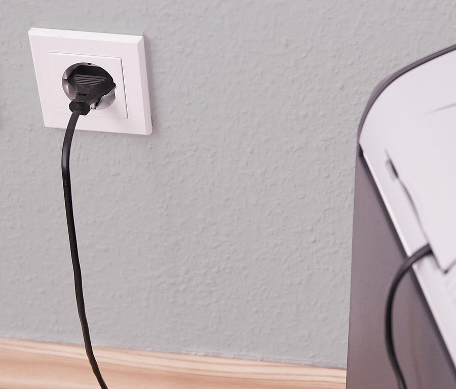 Schwarzer Stecker in weißer Steckdose an einer hellgrauen Wand mit einer Holzleiste am unteren Rand, das Kabel führt zu einer weißen und dunkelgrauen AEG Kühlbox »KK 28«.