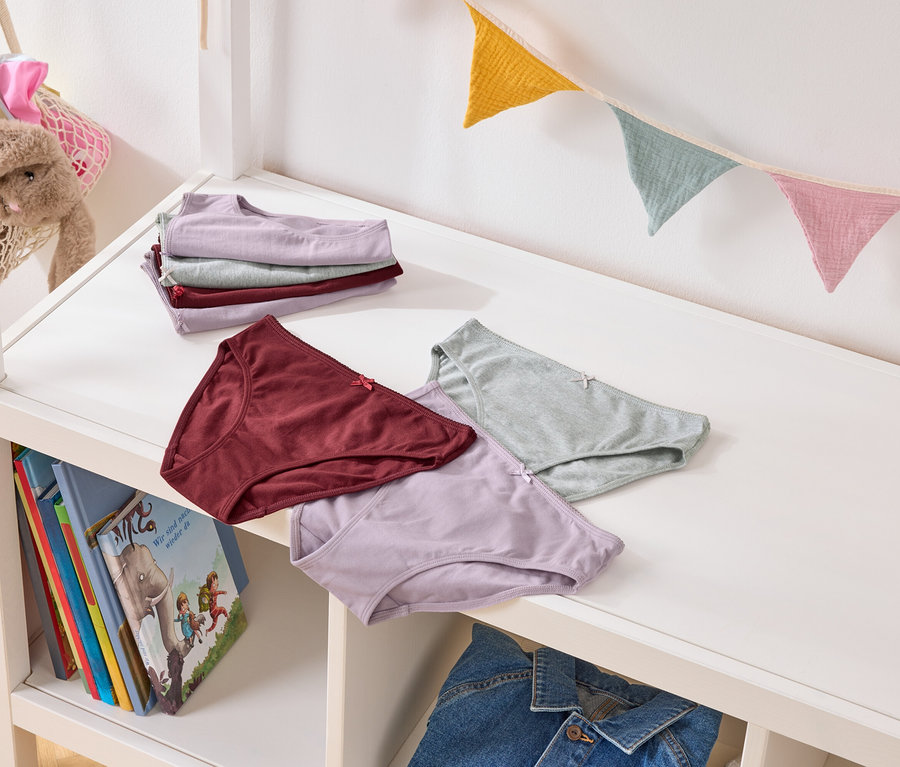 Mehrere Unterhosen in Pastellfarben liegen auf einem weißen Regal, darunter Bücher und eine Jeansjacke. Im Hintergrund hängt eine bunte Wimpelkette.