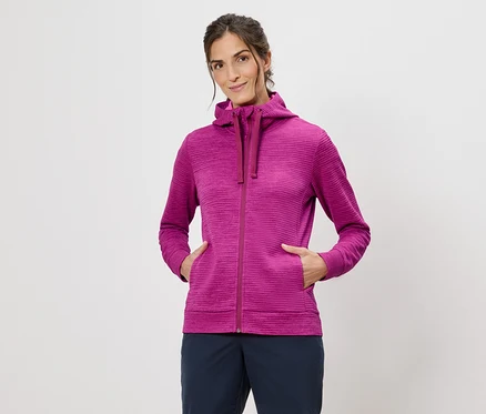 Frau mit Kapuzenpullover in Pink mit Händen in den Taschen vor einem weißen Hintergrund.