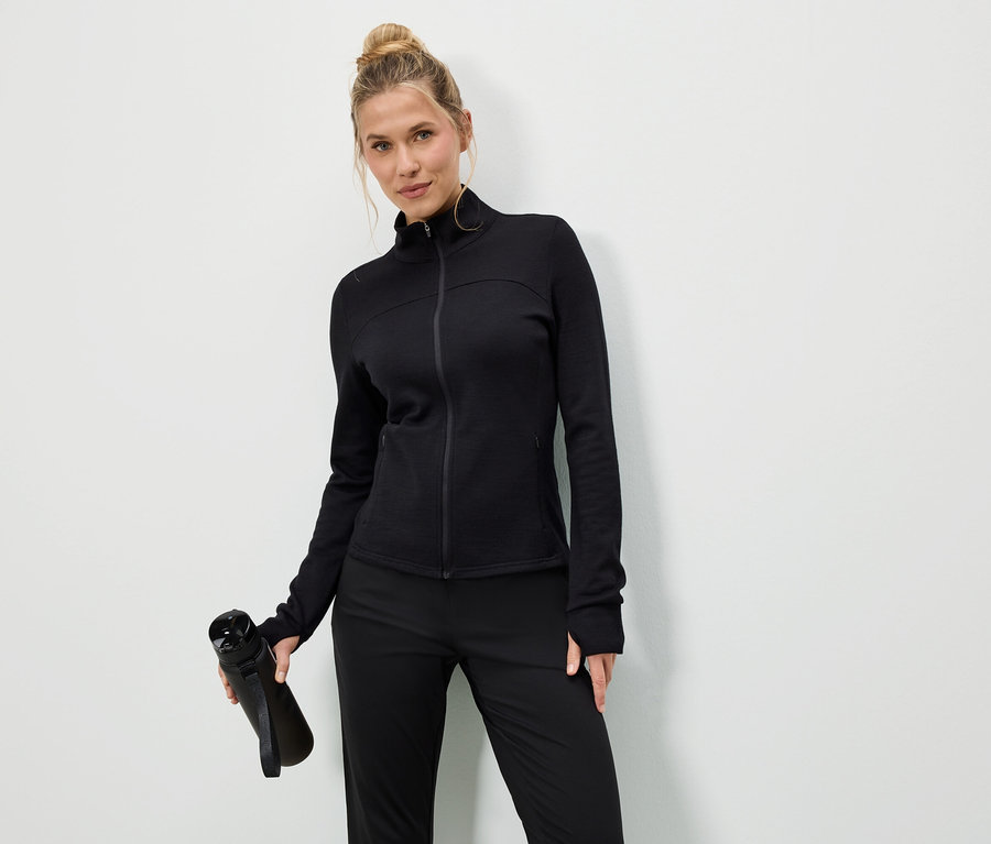 Frau in schwarzer Jacke und Hose hält eine schwarze Wasserflasche vor einem weißen Hintergrund.