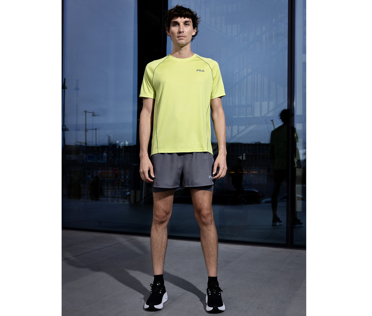 Mann steht vor Glasfassade, trägt ein hellgrünes FILA Herren-Funktionsshirt, graue Shorts und schwarze Sportschuhe.