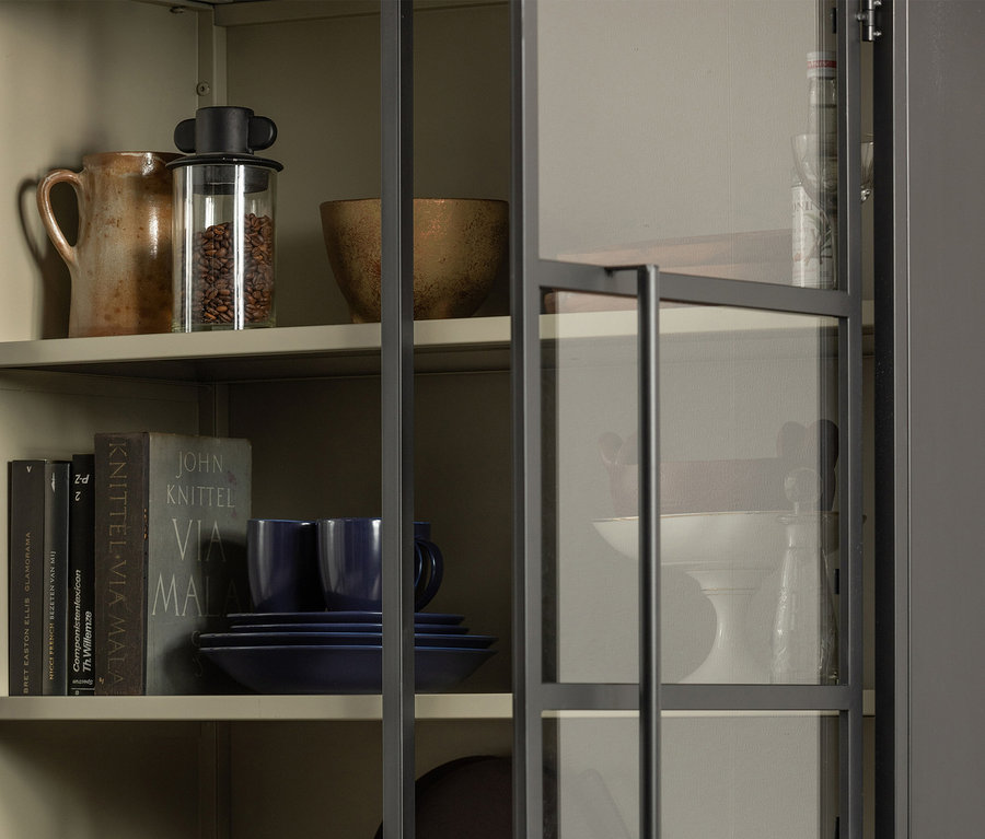 Hellbeiger Schrank mit dunkelgrau gerahmten Glastüren, der Gegenstände auf Regalen zeigt. Oberstes Regal: Keramikkrug, Glasgefäß mit Kaffeebohnen, bronzefarbene Schale. Mittleres Regal: Bücher, dunkelblaue Teller und Tassen. Unterstes Regal (teilweise sichtbar): weißer Tortenständer, Glasdekanter.