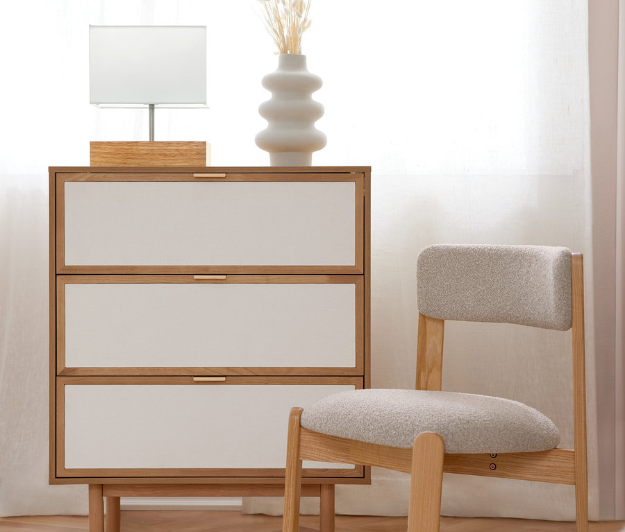Eine helle Holzkommode mit drei weißen Schubladen und goldenen Griffen, darauf eine weiße Tischlampe und eine weiße Vase mit Trockenblumen. Daneben steht ein heller Holzstuhl mit grau gepolsterter Sitzfläche. Der Raum hat einen Holzboden mit einem weißen Teppich und ein helles Fenster mit weißen Vorhängen.