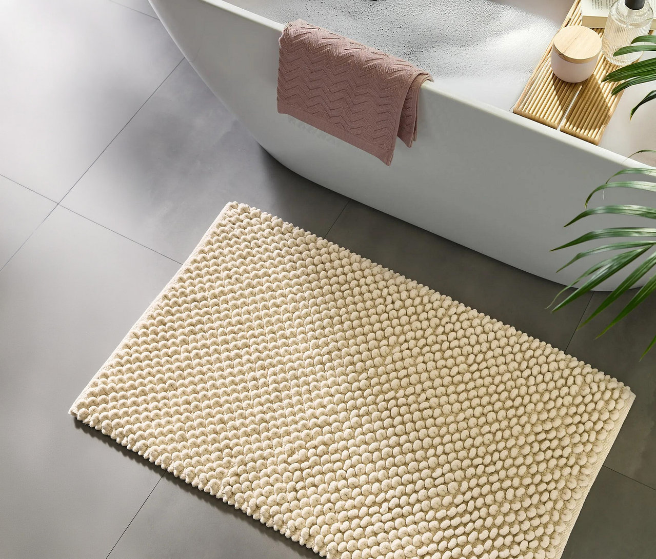 Ein cremefarbener Badteppich mit Noppenstruktur liegt auf einem grauen Fliesenboden neben einer weißen Badewanne. Ein rosafarbenes Handtuch hängt über dem Wannenrand, daneben ein Holztablett mit Pflegeprodukten und einem Buch. Eine grüne Pflanze ist rechts sichtbar.