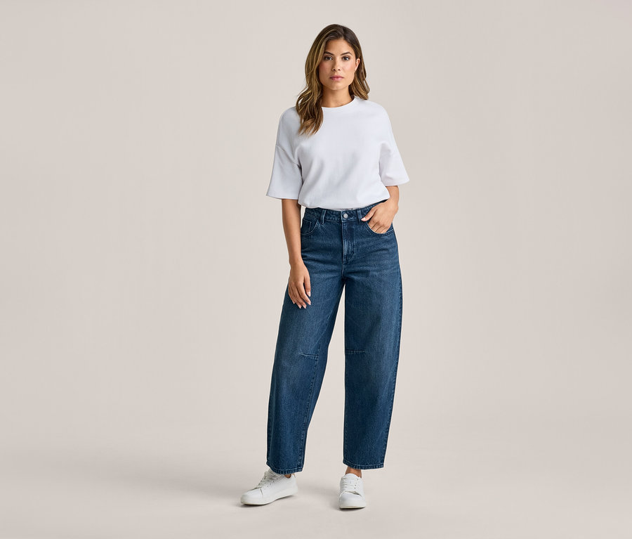 Frau steht, trägt ein weißes T-Shirt, dunkelblaue weite Jeans und weiße Sneaker. Ihre rechte Hand steckt in der Hosentasche.