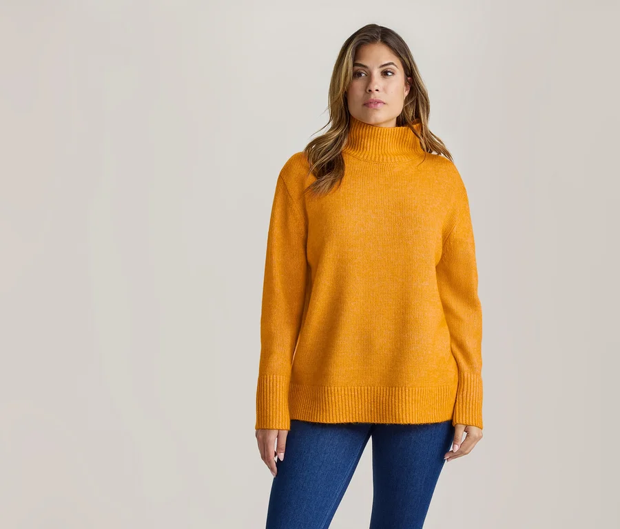 Eine Frau in einem orangefarbenen Rollkragenpullover und blauen Jeans steht vor hellem Hintergrund und blickt leicht nach rechts.