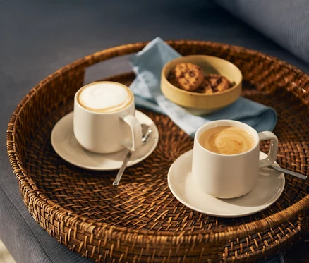 Zwei helle Kaffeetassen, eine mit Milchschaum und eine mit Latte-Art-Wirbel, stehen auf passenden Untertassen mit Löffeln auf einem runden geflochtenen Tablett. Eine kleine Schale mit Keksen und eine hellblaue Serviette sind ebenfalls auf dem Tablett.