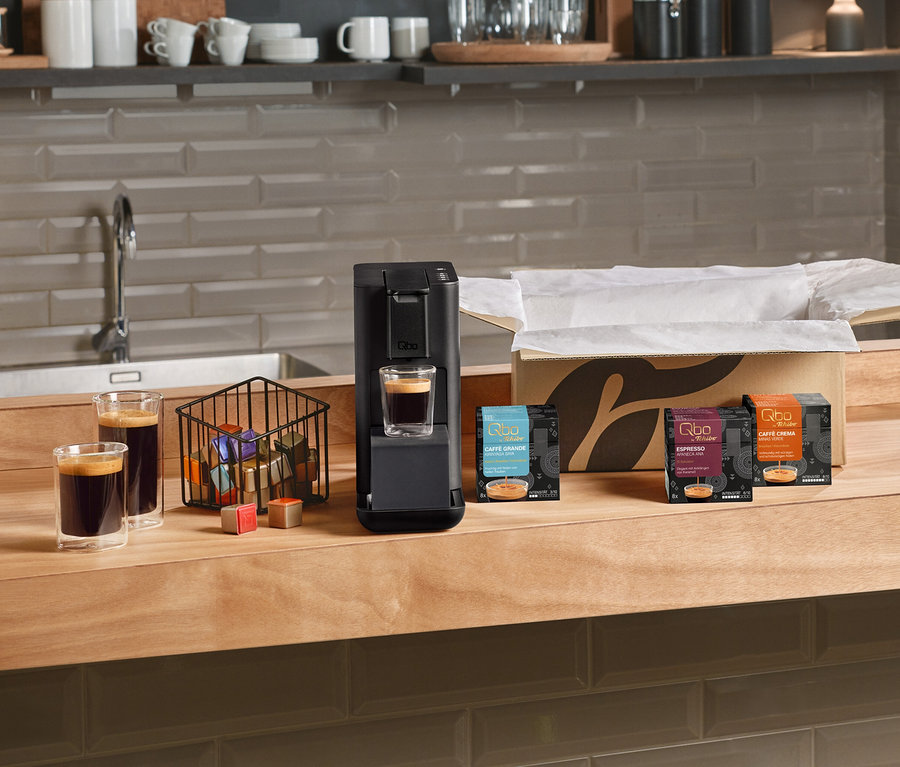 Die Kaffeemaschine Qbo Iconic, Dark Chrome 1,1 DAEE brüht Kaffee in ein Glas, daneben stehen zwei weitere Kaffeegläser, ein Korb mit bunten Kapseln und Packungen von Qbo Caffè Grande KIRINYAGA BAYA, Qbo Espresso APANECA ANA und Qbo Caffè MINAS VERDE auf einer Holztheke.