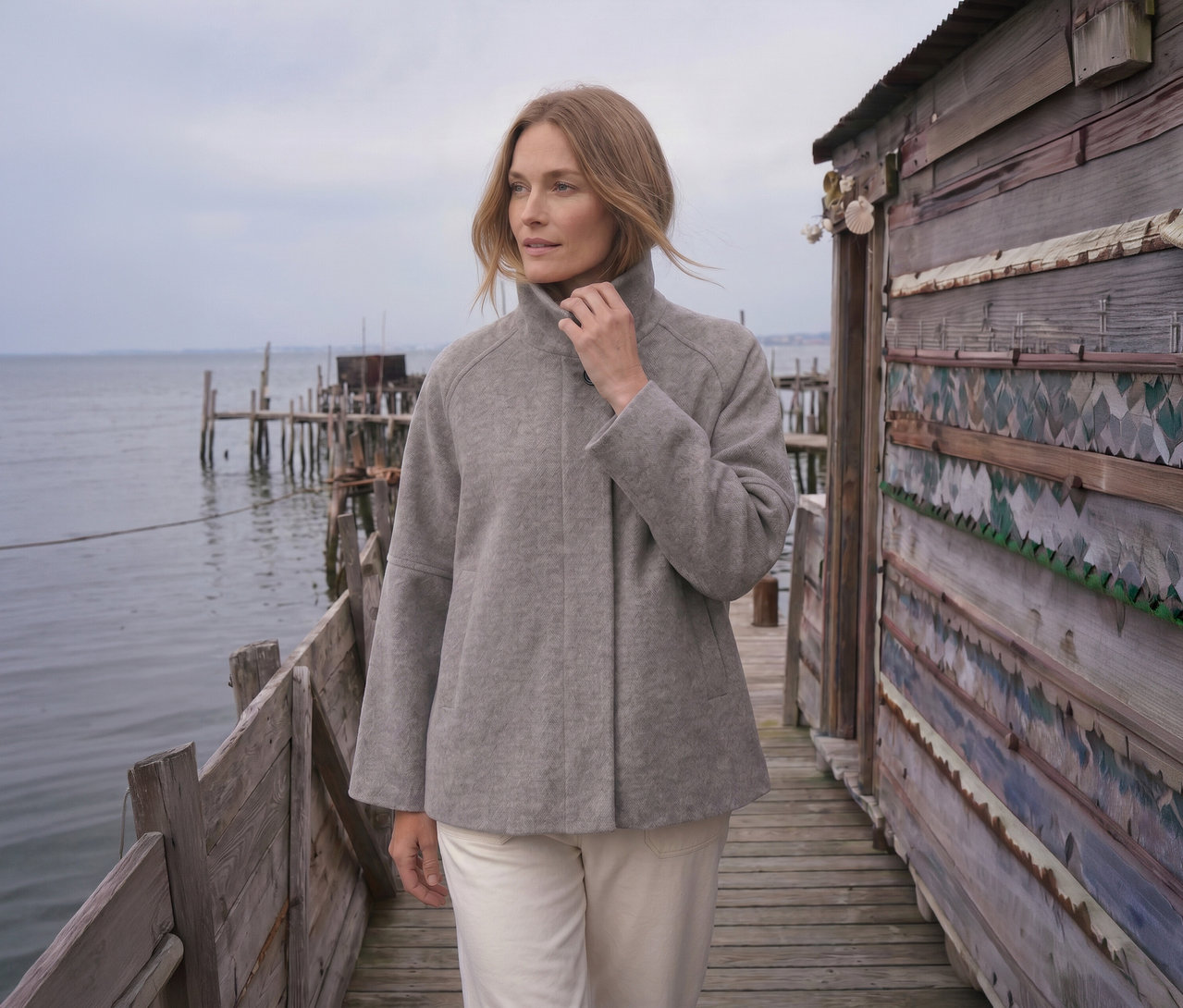 Frau in hellgrauer Jacke und cremefarbener Hose steht auf einem Holzsteg und blickt aufs Wasser. Ihre Hand ist am Jackenkragen, rechts ist eine rustikale Holzhütte.