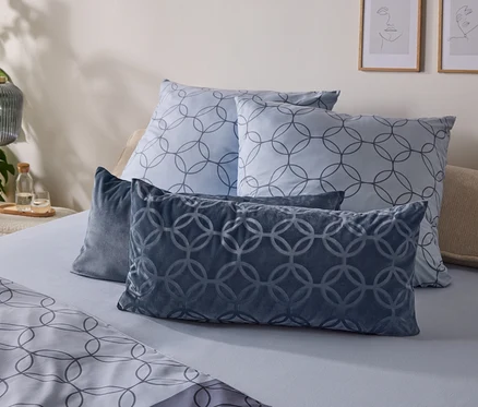 Ein Bett mit hellblauer Bettwäsche und zwei quadratischen Kissen mit grauem, ineinandergreifendem Kreismuster. Davor liegen zwei dunkelblaue Samtkissen mit erhabenem Kreismuster. Eine Pflanze, eine gerippte Glaslampe und gerahmte Linienkunst sind im Hintergrund zu sehen.