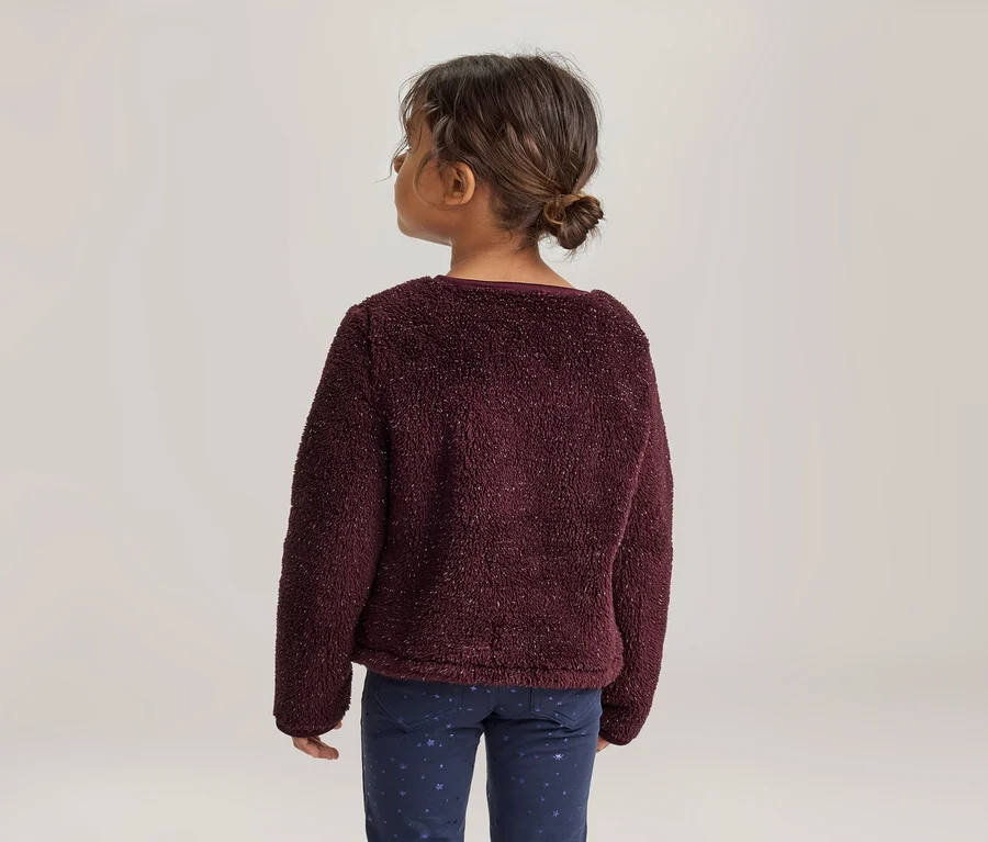 Junges Mädchen von hinten, trägt einen bordeauxroten flauschigen Pullover und dunkelblaue Hosen mit Sternenmuster, die Haare zu einem Dutt gebunden, blickt nach links.