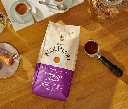 Auf einem Holztisch liegen eine Packung Caffè Molinari Espresso Perfetto-Bohnen, eine Tasse Espresso und Gebäckstücke.
