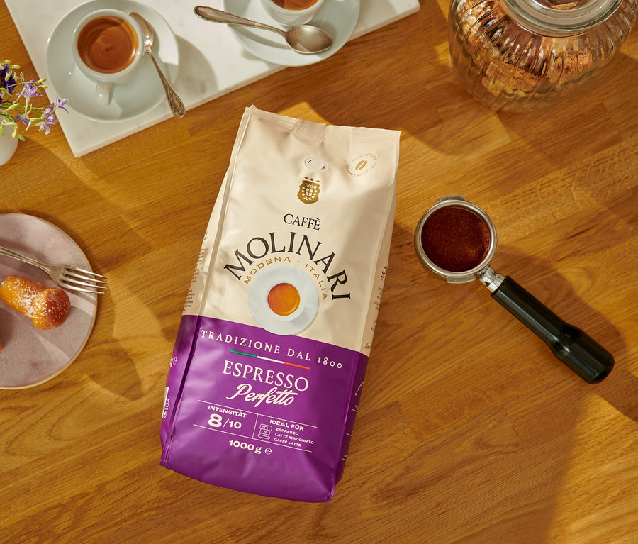Auf einem Holztisch liegen eine Packung Caffè Molinari Espresso Perfetto-Bohnen, eine Tasse Espresso und Gebäckstücke.